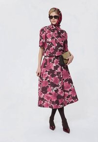 Femme portant une robe fleuri rose et rouge foncé avec un foulard assorti, des collants noirs, des talons rouge foncé, des lunettes de soleil et tenant une pochette beige.