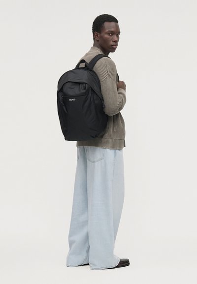 Un homme portant un pull couleur taupe et un jean à jambes larges présente un sac à dos noir HUGO sur un fond uni.