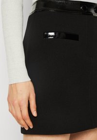 Gros plan sur une personne portant une jupe noire avec une ceinture brillante et des bordures de poche noires, associée à un haut léger à manches longues.