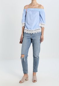 Blouse rayée bleu et blanc, épaules dénudées, avec bordure en dentelle, associée à un jean délavé bleu clair et des mules taupe à talons carrés.