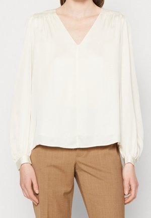 Woman wearing a long-sleeve, cream-colored V-neck blouse paired with light brown trousers, standing against a plain background.