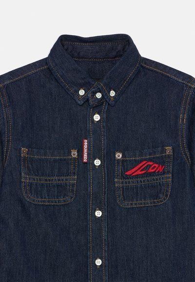 Chemise en denim foncé avec un col boutonné, dotée de deux poches avant, de boutons blancs et d'un logo brodé rouge sur la poche gauche.