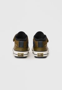 Hnědé plátěné vysoké tenisky s černým pruhem, suchým zipem a bílou gumovou podrážkou, se znakem Converse All Star na patě.