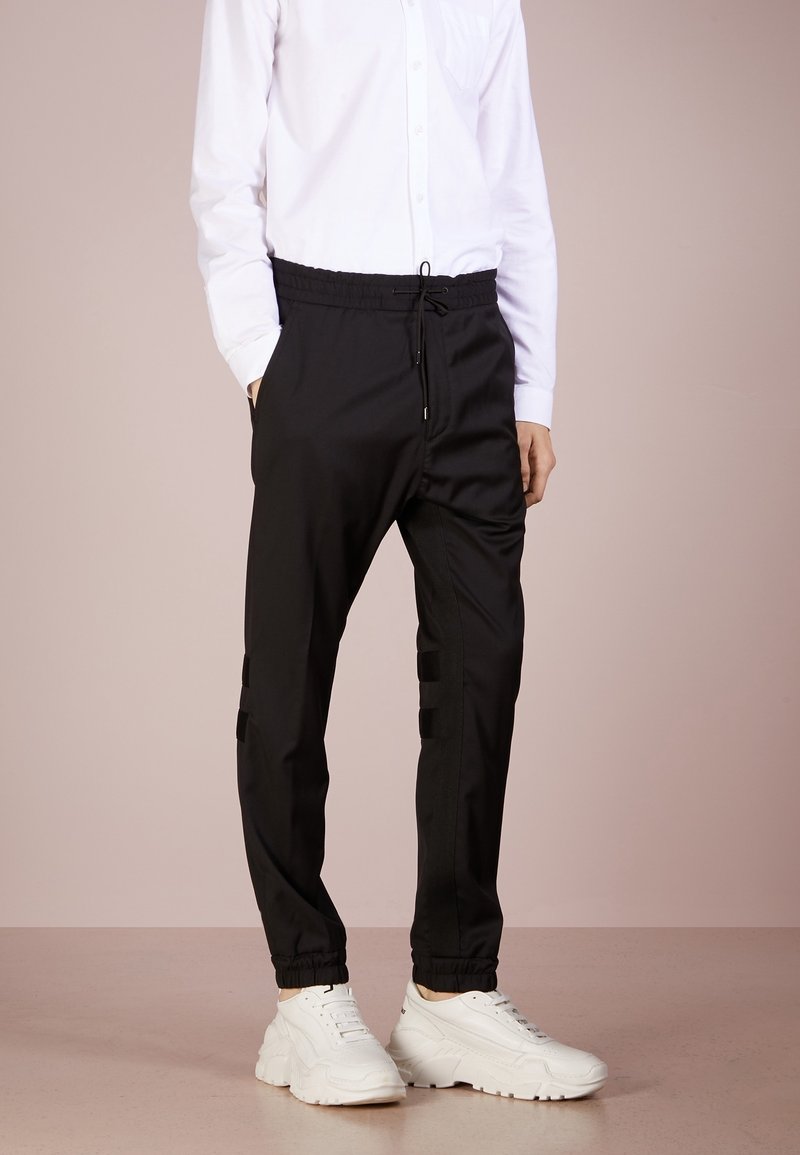 Person trägt schwarze Jogginghose mit Kordelzug und elastischen Bündchen, weißes langärmeliges Hemd mit Knöpfen und weiße klobige Sneakers auf beigem Boden.