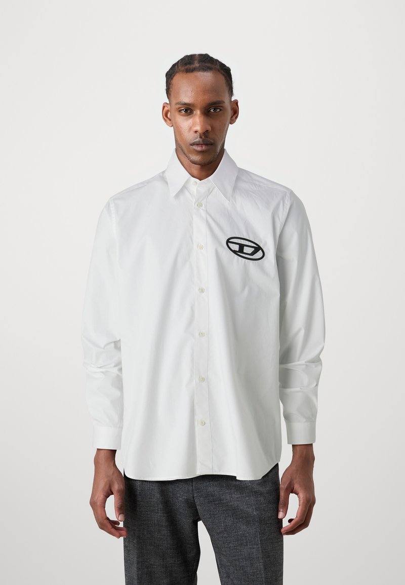 Chemise blanche à boutons en tissu robuste avec un col pointu et un logo noir brodé sur la poitrine. Manches longues, coupe légèrement décontractée.