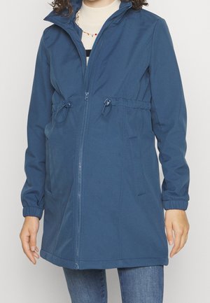 Femme portant une veste bleue à fermeture éclair avec des cordons de taille ajustables et des poignets élastiques, associée à un jean bleu et un haut blanc en dessous.