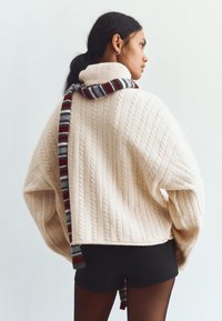 Kremowy sweter w warkoczowe sploty z wysokim kołnierzem, w zestawieniu z wielokolorowym, pasiastym szalem. Sylwetka jest luźna i krótsza.