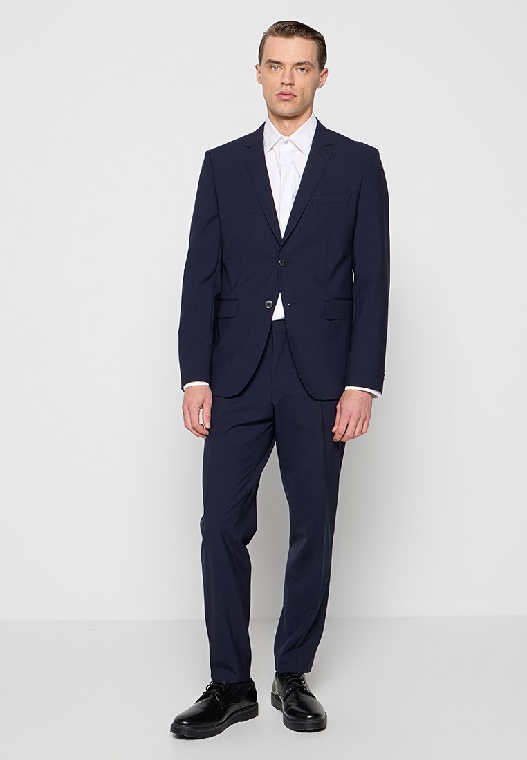 Mann, der vor einem weißen Hintergrund steht, trägt einen marineblauen Anzug, ein weißes Hemd und schwarze Anzugschuhe, steht mit entspannten Armen nach vorne gerichtet.