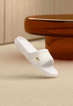 Sandalo tipo ciabatta Lacoste in bianco e azzurro chiaro. Presenta una fascia testurizzata con il logo, suola piatta e un design minimalista su una superficie morbida.