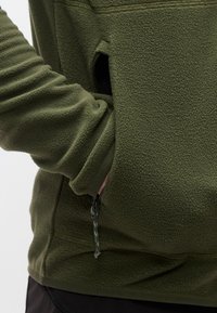 Άτομο που φοράει φούτερ από φλις ελιάς, με το χέρι μέσα στην τσέπη στο πλάι με φερμουάρ, δείχνοντας την υφή και τη λεπτομέρεια του φερμουάρ.