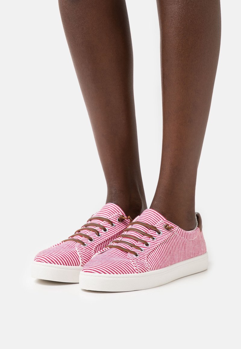 Pieds portant des baskets basses rayées rouges et blanches avec des lacets marron et des semelles blanches sur un fond clair uni.