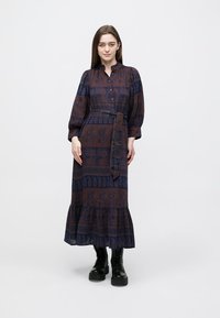 Long, patterned dress in dark blue and brown with a cinched waist, button front, puffed sleeves, and a tiered skirt. Paired with black boots.