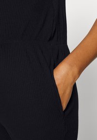 Macacão preto com textura em tecido canelado, incluindo um detalhe de bolso lateral, que destaca a sua textura e silhueta ajustada. Acabamento suave e design elástico.