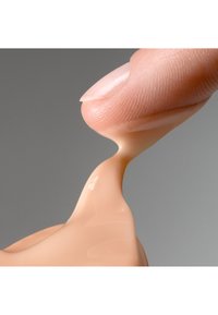 Fondotinta liquido in una tonalità pesca-beige, che scorre dalla punta di un dito su uno sfondo grigio chiaro e liscio. La texture appare cremosa e lucida.
