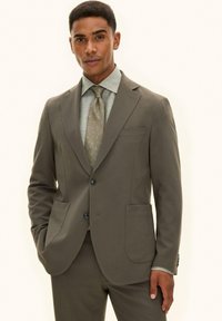 Mężczyzna ubrany w oliwkowo-zielony garnitur na dwa guziki, z pasującymi spodniami, jasnooliwianą koszulą i wzorzystym krawatem, stojący z jedną ręką w kieszeni.