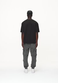 Mies, który nosi czarną czapkę, czarną oversize'ową koszulę, szare spodnie cargo i białe trampki, stoi tyłem na białym tle.