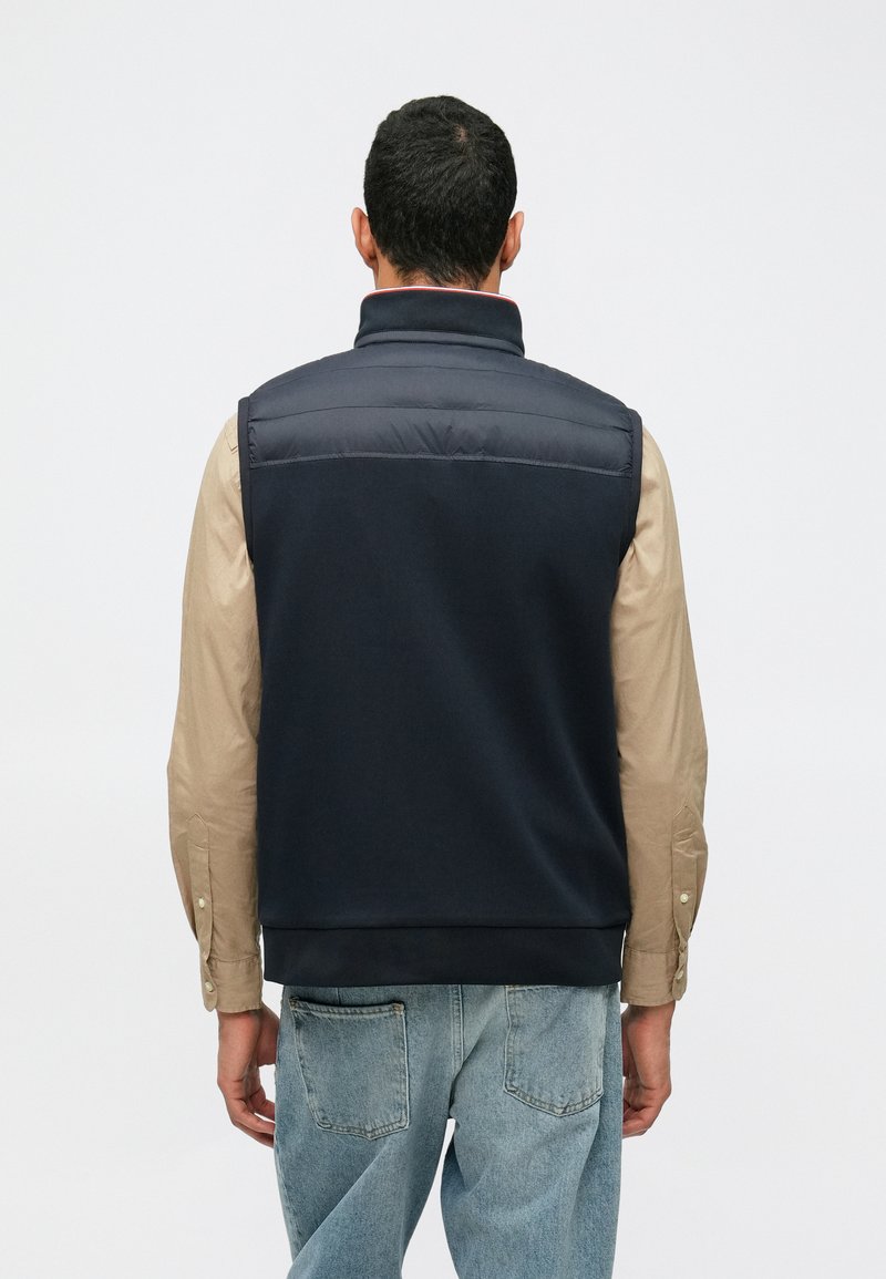 Marineblaues ärmelloses Weste mit einem gesteppten oberen Abschnitt, glattem Stoff und elastischem Saum. Darunter ein hellbeiges Langarmshirt.