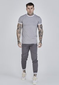 Grå kortærmet t-shirt med hvide detaljer, prydet med "SIKSILK"-logo, parret med mørkegrå tætsiddende joggers og hvide sneakers.