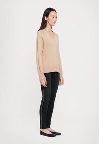 Suéter beige de cuello en V con puños y dobladillo acanalados, combinado con jeans negros que presentan un detalle de cremallera lateral. La modelo lleva zapatos planos negros.