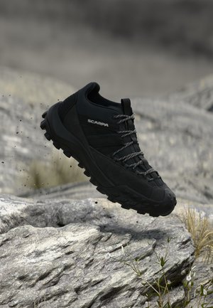 Schwarzer Scarpa Wanderschuh mit robuster Sohle scheint über felsigem Gelände mit kleinen Pflanzen und unscharfem Hintergrund zu schweben.