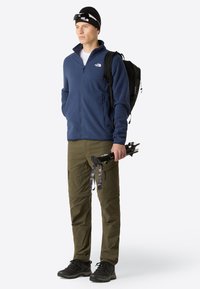 Giacca in pile blu da uomo, pantaloni cargo oliva, zaino nero, che tiene bastoni da trekking neri. L'outfit presenta tasche con zip e materiali testurizzati.