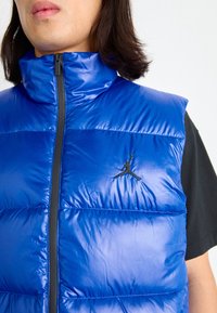 Blå vadderad väst med hög krage, med en svart dragkedja och en liten pr äglad logotyp. Materialet har en glänsande, slät textur.