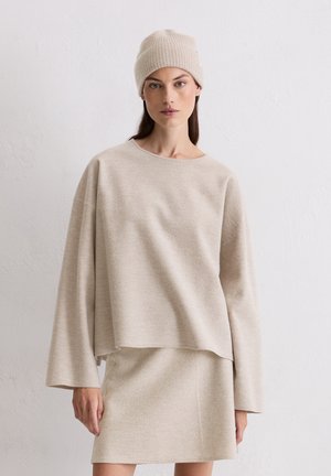 Beige gestrickter Pullover mit langen, weiten Ärmeln, kombiniert mit einem passenden, taillierten Rock. Das Material sieht weich und strukturiert aus. Das Model trägt eine gerippte M�ütze.