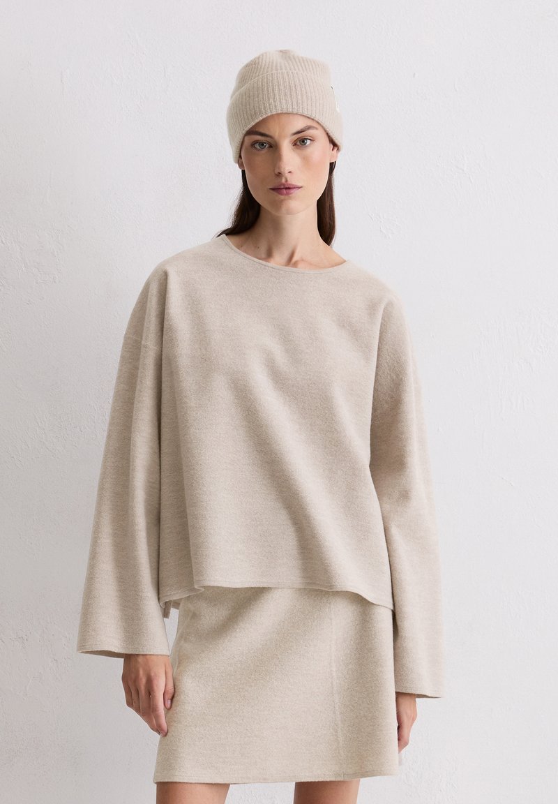Beżowy sweter dziany z długimi, szerokimi rękawami, w zestawie z dopasowaną spódnicą. Materiał wydaje się miękki i teksturowany. Modelka nosi prążkowaną czapkę.