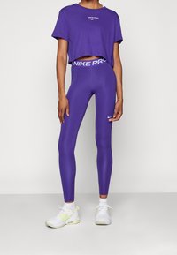 Fioletowa krótka koszulka z logo "NIKE PRO", połączona z fioletowymi legginsami o kontrastowym pasie. Uzupełniona białymi butami sportowymi.