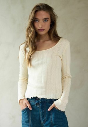 Világos krém színű, hosszú ujjú felső bordázott textúrával, kerek nyakkivágással, fodros aljjal és finom mintázattal. Farmerral kombinálva.