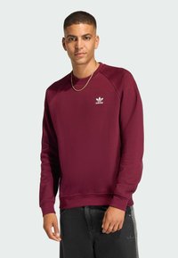 Burgundy sweatshirt made of cotton blend, featuring a ribbed crew neck and cuffs. White adidas logo on the chest. Black jeans with side stripes.