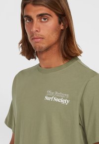 Camiseta de algodón verde oliva con cuello redondo, que presenta texto blanco: "La Sociedad de Surf del Futuro" en el frontal. Mangas cortas, corte relajado.