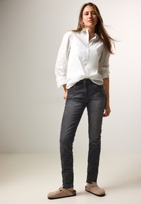 Camisa blanca de botones con mangas enrolladas, combinada con unos jeans de mezclilla oscuros de corte ajustado y un sutil efecto desgastado. Calzado ligero.