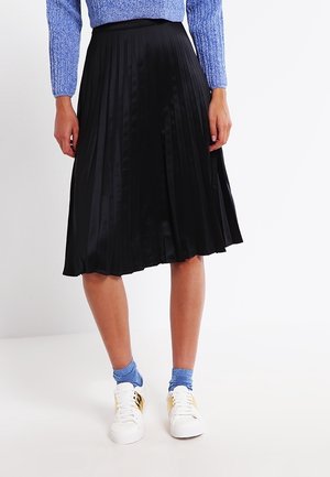 Schwarzer plissierter Midirock mit glatter Textur, getragen mit einem blauen Strickpullover und weißen Sneakern mit goldenen Akzenten.