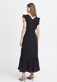 Robe maxi noire avec des manches courtes volantes, un dos en V et une jupe à volants. Tissu fluide avec une silhouette élégante, portée avec des talons verts.