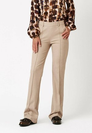 Woman wearing beige straight-leg trousers with front seams, a brown patterned long-sleeve blouse, and black loafers with fringe detailing.