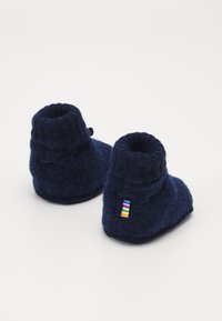Botitas de bebé de fieltro azul marino con un puño de punto, que cuentan con una pequeña etiqueta de rayas multicolores en el lateral. Textura suave y forma redondeada.