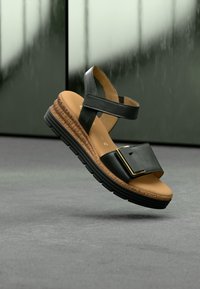 Zwarte leren sandaal met twee verstelbare banden, een platte zool en een textuurachtige, gevlochten middenzool. Goudkleurige beslagaccenten op de voorste band.