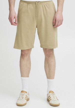 Shorts en tricot beige avec une taille élastique et un cordon de serrage. Comprend des poches latérales et une coupe décontractée, atteignant mi-cuisse.