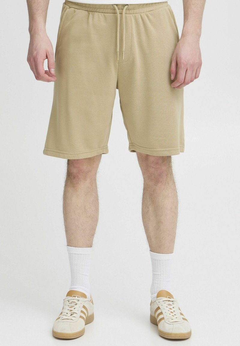 Shorts en tricot beige avec une taille élastique et un cordon de serrage. Comprend des poches latérales et une coupe décontractée, atteignant mi-cuisse.