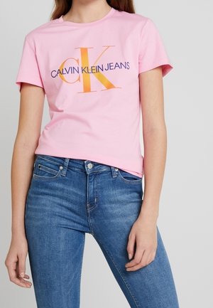 Woman wearing a pink Calvin Klein Jeans t-shirt with logo and fitted blue jeans, standing against a plain light gray background.
