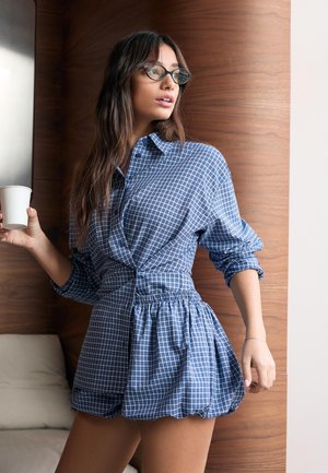 Mlada ženska v modri obleki s karo vzorcem in očali, ki drži belo skodelico, stoji v prostoru ob leseni steni.