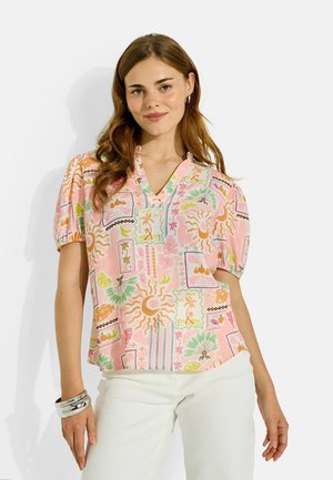 Jeune femme aux longs cheveux bruns portant un chemisier coloré à manches courtes à motifs et un pantalon blanc, souriante, les mains détendues sur les côtés.