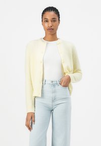 Cardigan lavorato a maglia di un giallo chiaro con scollatura rotonda e chiusura con bottoni. Indossato su una maglietta bianca aderente, abbinato a jeans alta vita di un azzurro chiaro.