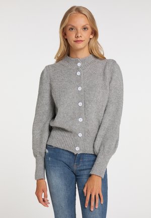 Junge Frau trägt eine graue, geknöpfte Strickjacke mit Puffärmeln und blaue Jeans, steht vor einem schlichten Hintergrund.