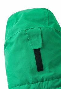 Grüne wasserdichte Jacke in der Ecke mit glatter Textur, schwarzem Klettverschluss und passgenauer Form mit minimalen Nähdetails.