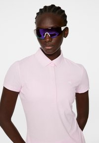 Różowa koszulka polo z fakturowanej tkaniny, krótkie rękawy i kołnierz. Noszona z ciemnymi, sportowymi okularami przeciwsłonecznymi z fioletowymi soczewkami.