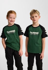 Två barn bär grön Hummel sporttröja med kort ärm och svarta raglansleeves. Båda har svarta byxor. Tröjorna har vita logotyper.