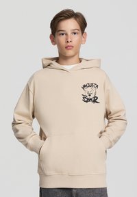 Felpa beige con tasca anteriore, cappuccio con lacci e grafica nera che presenta un orso e il testo "SONO SOLO UN ORSO". Materiale morbido e vestibilità comoda.