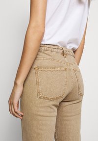 Jeans de talle alto en beige claro con una silueta ajustada, costuras visibles, dos bolsillos traseros y una tela texturizada.